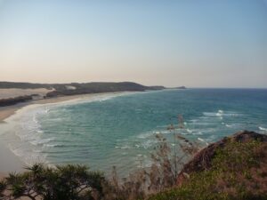 A wide sandy beach curves along the Fraser Island coastline, with gentle waves, clear blue-green water, and sparse vegetation on the rocky foreground under a clear sky.