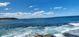 Palm Beach View of a clear blue ocean with gentle waves and foam, distant shoreline, and a bright sky scattered with small white clouds. google map photo
