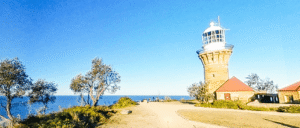 A stone lighthouse with a glass lantern room stands near red-roofed buildings by the sea in Palm Beach, under a clear blue sky, with trees and a dirt path in the foreground.