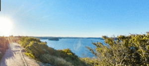 A bright sun shines over Palm Beachs coastal path lined with bushes, overlooking a calm blue body of water with distant land visible on the horizon under a clear blue sky.