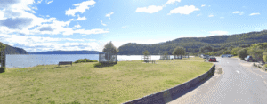 A grassy park area with benches and picnic tables overlooks a calm body of water at Palm Beach, surrounded by tree-covered hills. A road with parked cars runs along the right side under a bright, partly cloudy sky.